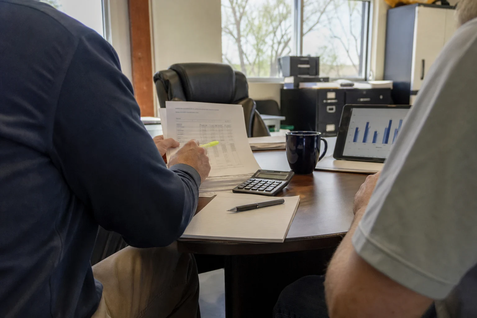 Two men at a round office table reviewing a printed statement with a highlighter, beside a calculator, notepad, pen, coffee mug and tablet showing a bar chart