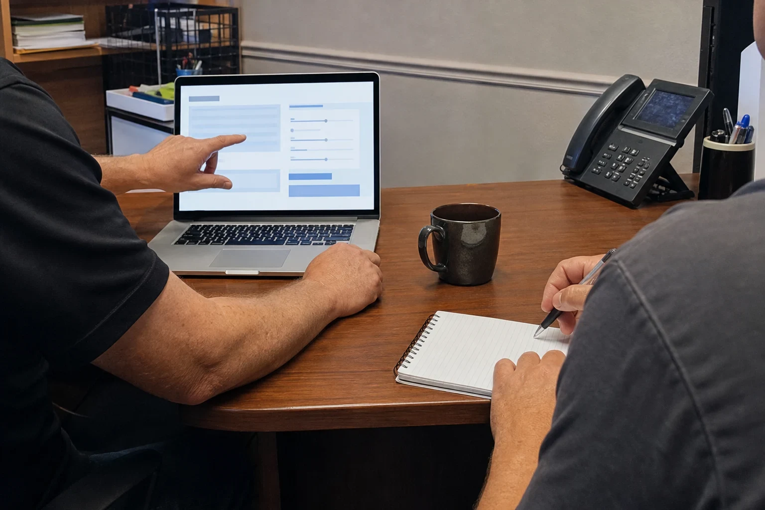 Agent points to adjustable slider options on a laptop at a wooden office desk while a client takes notes in a spiral notebook beside a desk phone and mug