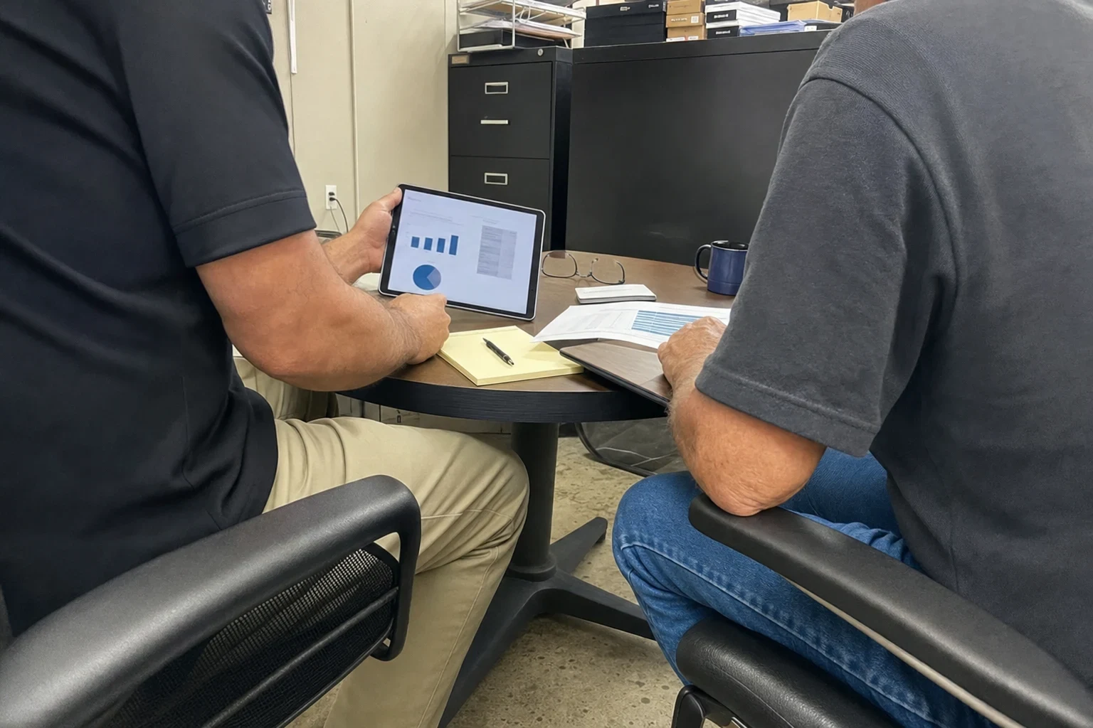 Two men seated at a small round table in a back office, one holding a tablet showing charts while the other reviews a printed document, with filing cabinets behind.